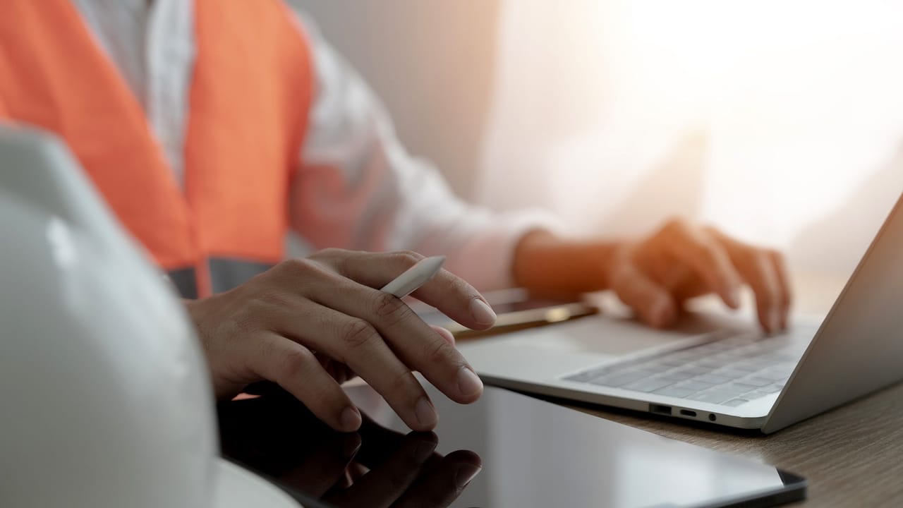 Close-up of a project engineer working on a laptop with a hard hat on the desk beside them