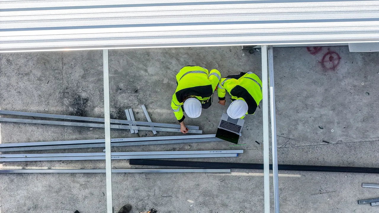 Overhead view of two workers raising an inspection request on a tablet beside stacked steel framing